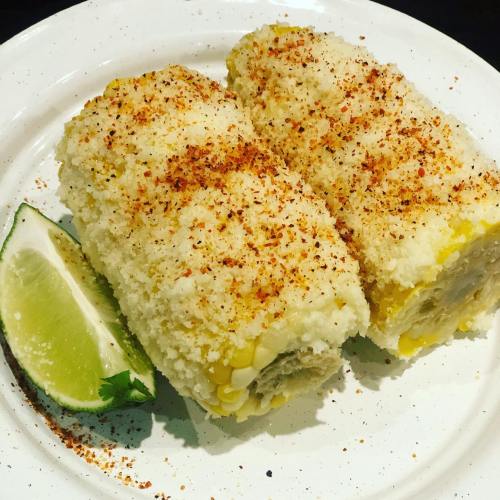 Two elote corn on the cob pieces, coated in cheese and spices, served with a lime wedge on a white plate.