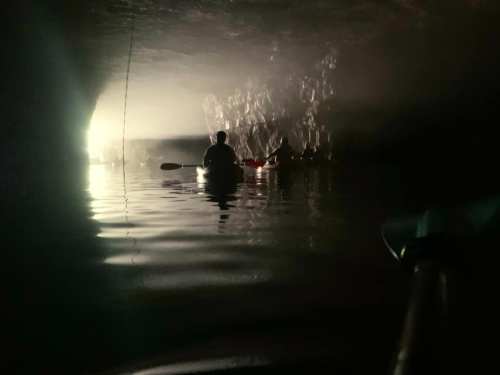 Two kayakers explore a dimly lit cave, with reflections on the water and light illuminating the rocky walls.