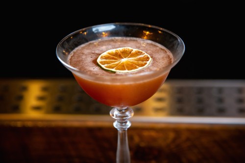 A cocktail in a elegant glass, garnished with a dried lime slice, resting on a bar counter.