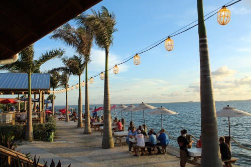 A waterfront scene with palm trees, string lights, and people dining at outdoor tables by the water during sunset.
