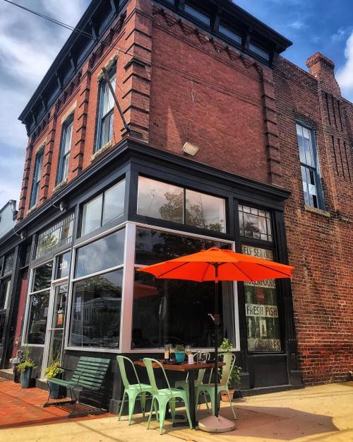 A corner building with large windows, featuring an orange umbrella over a table and green chairs outside.