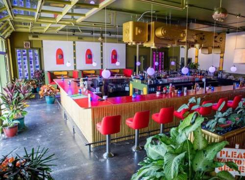 Vibrant bar interior with colorful seating, plants, and a stylish counter, featuring retro decor and ambient lighting.