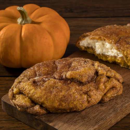 A pumpkin and a freshly baked pumpkin cookie on a wooden surface, with a slice revealing creamy filling.