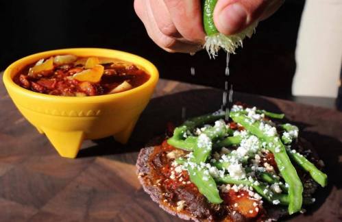A hand squeezes lime over a taco topped with green peppers and cheese, alongside a bowl of chili.