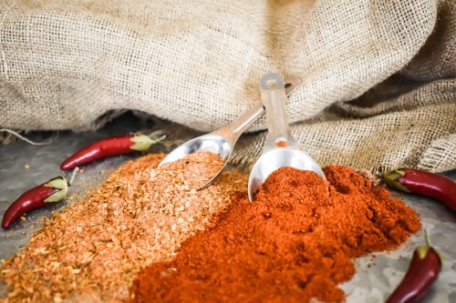Two metal scoops filled with red and brown spices, surrounded by red chili peppers on a textured surface with burlap.