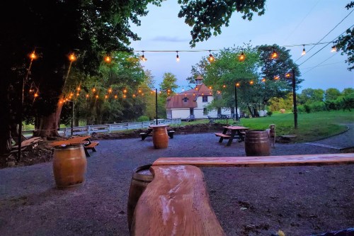 Outdoor seating area with string lights, wooden barrels, and a rustic house in the background, surrounded by trees.