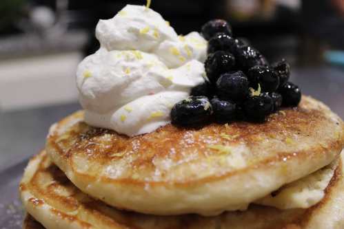 Stack of fluffy pancakes topped with whipped cream, blueberries, and a sprinkle of lemon zest.