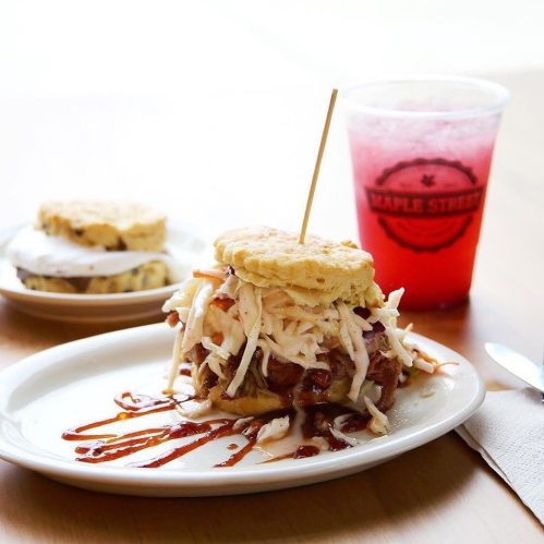 A pulled pork sandwich topped with coleslaw on a plate, alongside a refreshing drink and a biscuit dessert.