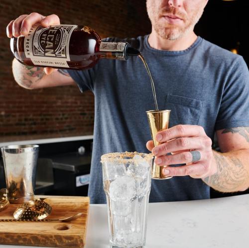 A person pours whiskey from a bottle into a jigger over a glass filled with ice, with bar tools in the background.