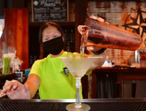 A bartender in a neon green shirt pours a drink into a martini glass with olives and a cherry.