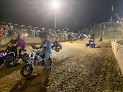 Children riding small electric vehicles on a dirt track at night, with a crowd watching in the background.