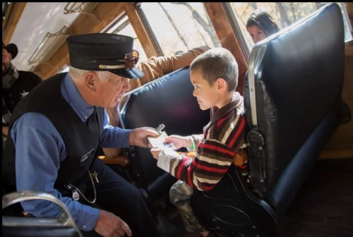 A train conductor interacts with a young boy seated on a train, both smiling and engaged in conversation.