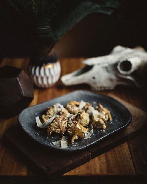 A dark plate of food featuring cauliflower, garnished with coconut and herbs, on a wooden table with decorative elements.