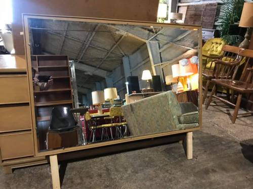 A large mirror reflecting furniture and lamps in a vintage shop setting.