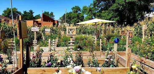 A vibrant garden with wooden planters, colorful flowers, and decorative art pieces, surrounded by greenery and a yellow umbrella.