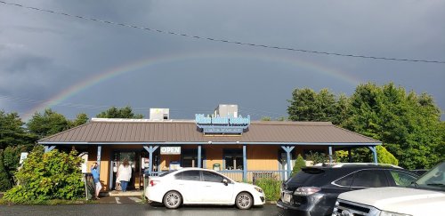 A restaurant with a rainbow in the sky above, surrounded by greenery and parked cars. Dark clouds loom in the background.