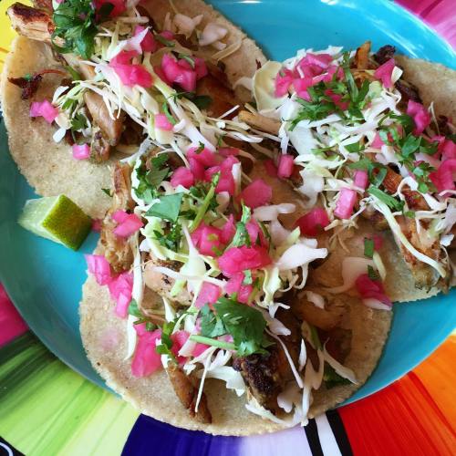 Three tacos filled with grilled meat, topped with cabbage, pink onions, cilantro, and a lime wedge on a colorful plate.