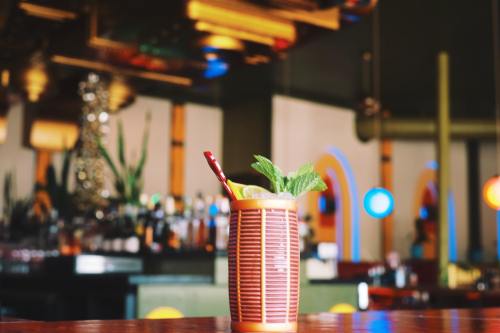 A colorful cocktail with mint and a lemon slice sits on a bar, with a blurred background of bottles and vibrant lights.
