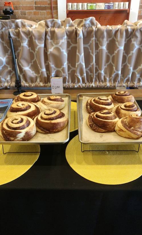 Two trays of freshly baked cinnamon rolls on a table, with a price tag of $6.00 displayed.