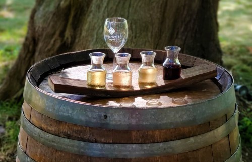 A wooden barrel holds a tray with four small glass carafes of different colored liquids and a wine glass.
