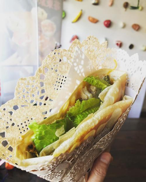 A hand holding a wrapped taco filled with lettuce and other ingredients, presented in a decorative lace paper.