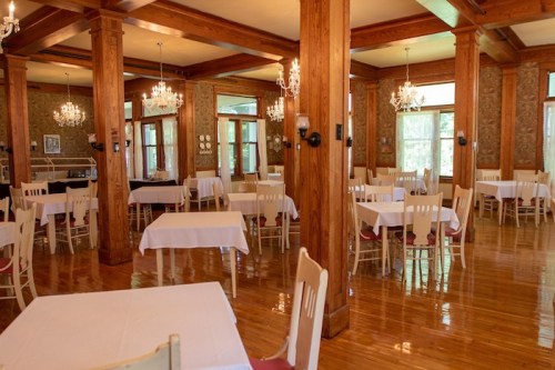 A spacious dining area with wooden pillars, white tablecloths, and chandeliers, featuring multiple empty tables.