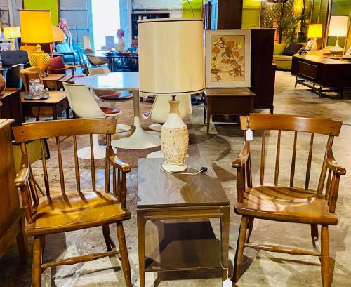 Two wooden chairs flanking a small table with a lamp, surrounded by vintage furniture in a bright, spacious room.