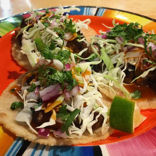 Three tacos topped with shredded cabbage, onions, cilantro, and sauce, served on a colorful plate with a lime wedge.