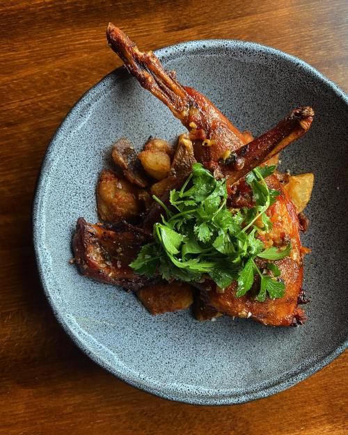 A plate of roasted meat with crispy potatoes, topped with fresh herbs, served in a textured bowl on a wooden table.