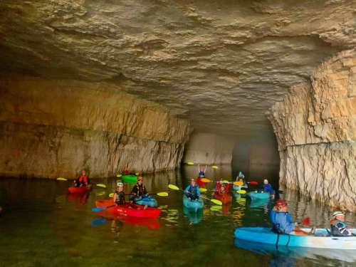 A group of kayakers in colorful kayaks paddles through a cave with rocky walls and calm water.