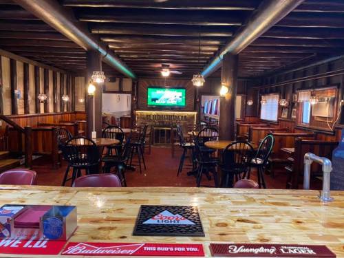 Cozy bar interior with wooden beams, tables, and a TV, featuring a polished wooden counter and various beer advertisements.