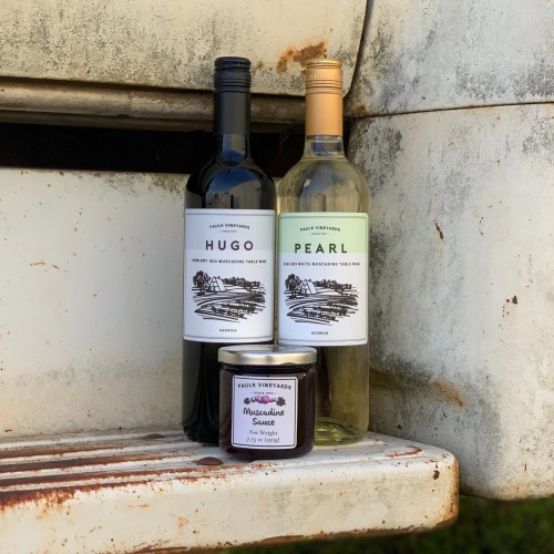 Two wine bottles labeled "Hugo" and "Pearl" alongside a jar of muscadine sauce on a weathered surface.