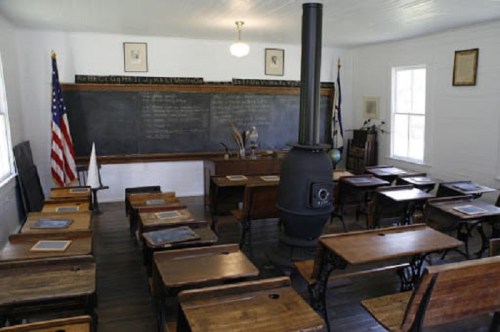 A vintage classroom with wooden desks, a blackboard, and a stove, featuring an American flag in the corner.