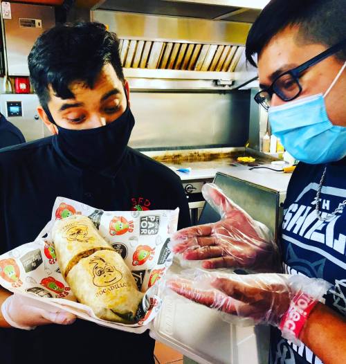 Two men in a kitchen, one wearing gloves and a mask, admiring a large burrito in a takeout container.