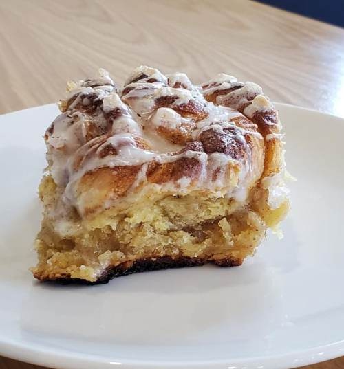 A slice of cinnamon roll cake with icing on a white plate, resting on a wooden table.