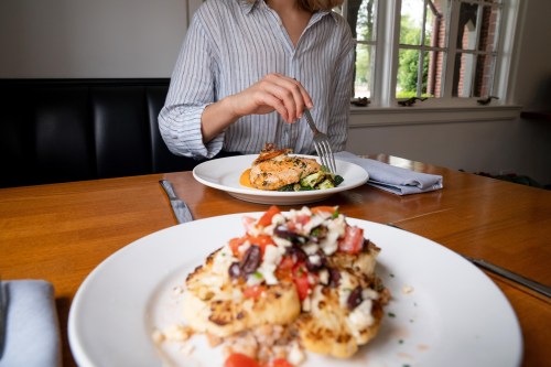 A person in a striped shirt enjoys a meal, featuring salmon and a colorful vegetable dish on a wooden table.