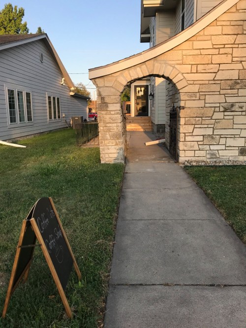 A stone archway leads to a pathway beside a grassy area, with a chalkboard sign for a coffee shop nearby.