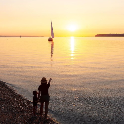 A silhouette of a person and a child by the water, watching a sailboat against a sunset backdrop.