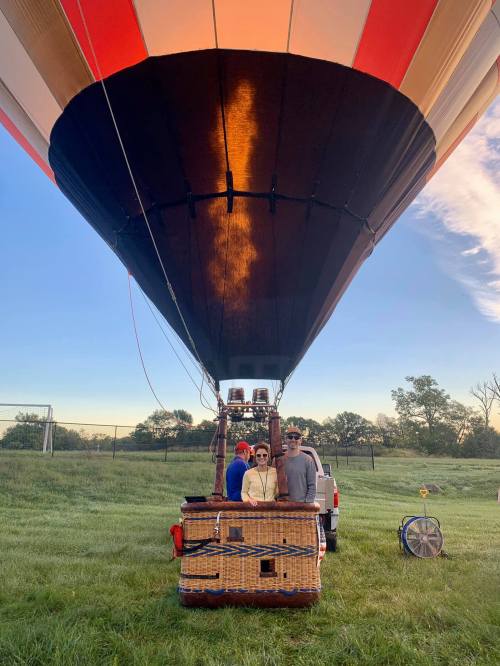 A hot air balloon on the ground with a wicker basket and two people standing in front, surrounded by grass and trees.