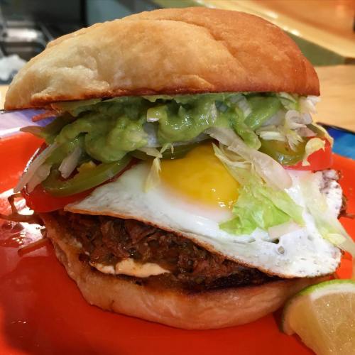 A delicious sandwich with pulled meat, a fried egg, lettuce, tomato, guacamole, and jalapeños on a toasted bun.