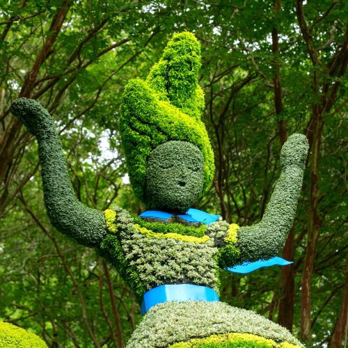 A topiary sculpture of a figure with green foliage, wearing a blue ribbon, surrounded by trees.