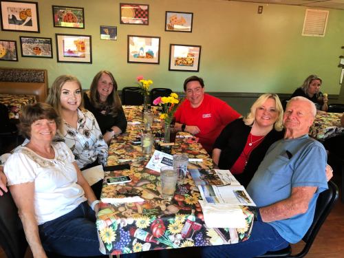 A group of six people sitting at a table in a restaurant, smiling and enjoying their time together. Colorful decor in the background.