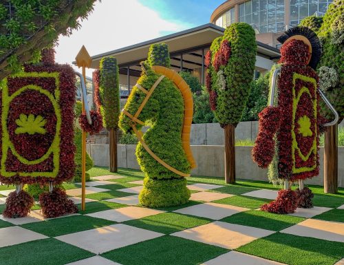 Colorful topiary figures resembling chess pieces in a garden setting, with a checkered pattern on the ground.