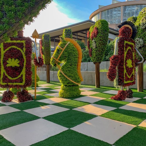 Colorful topiary figures resembling chess pieces in a garden setting, with a checkered green and white floor.