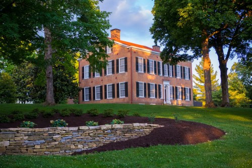 A historic brick house with black shutters, surrounded by lush greenery and a stone garden wall under a blue sky.