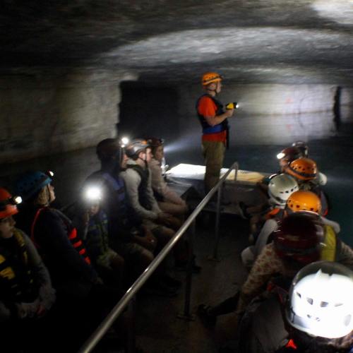 A group of people in helmets sits in a boat inside a dark cave, listening to a guide with a flashlight.