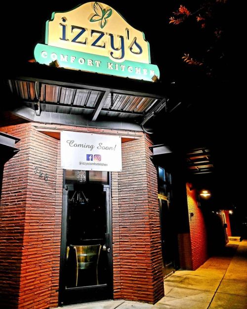 Sign for Izzy's Comfort Kitchen, with a "Coming Soon!" banner, illuminated at night. Red brick exterior.
