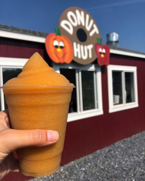 A person holds a large orange slushy drink in front of a red building with a "Donut Hut" sign featuring smiling pumpkins.