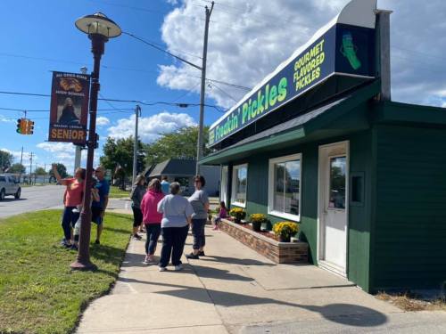 A group of people stands outside a green building with a sign reading "Ticklin' Pickles," a gourmet pickle shop.
