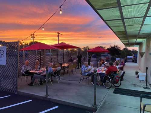 Outdoor dining scene at sunset, with patrons enjoying meals under red umbrellas and string lights.
